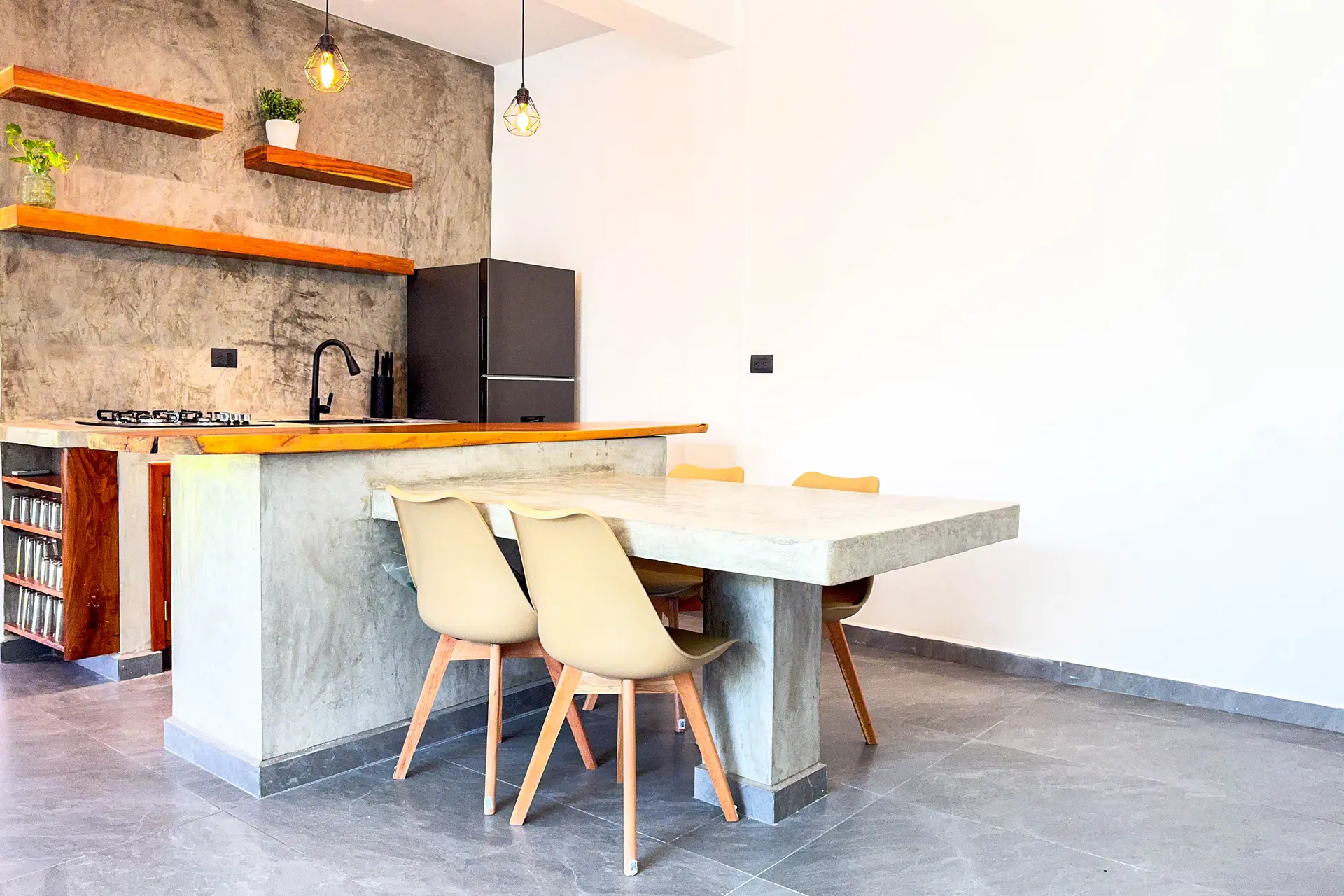 Cocina con barra de concreto y mesa integrada, repisas de madera y refrigerador