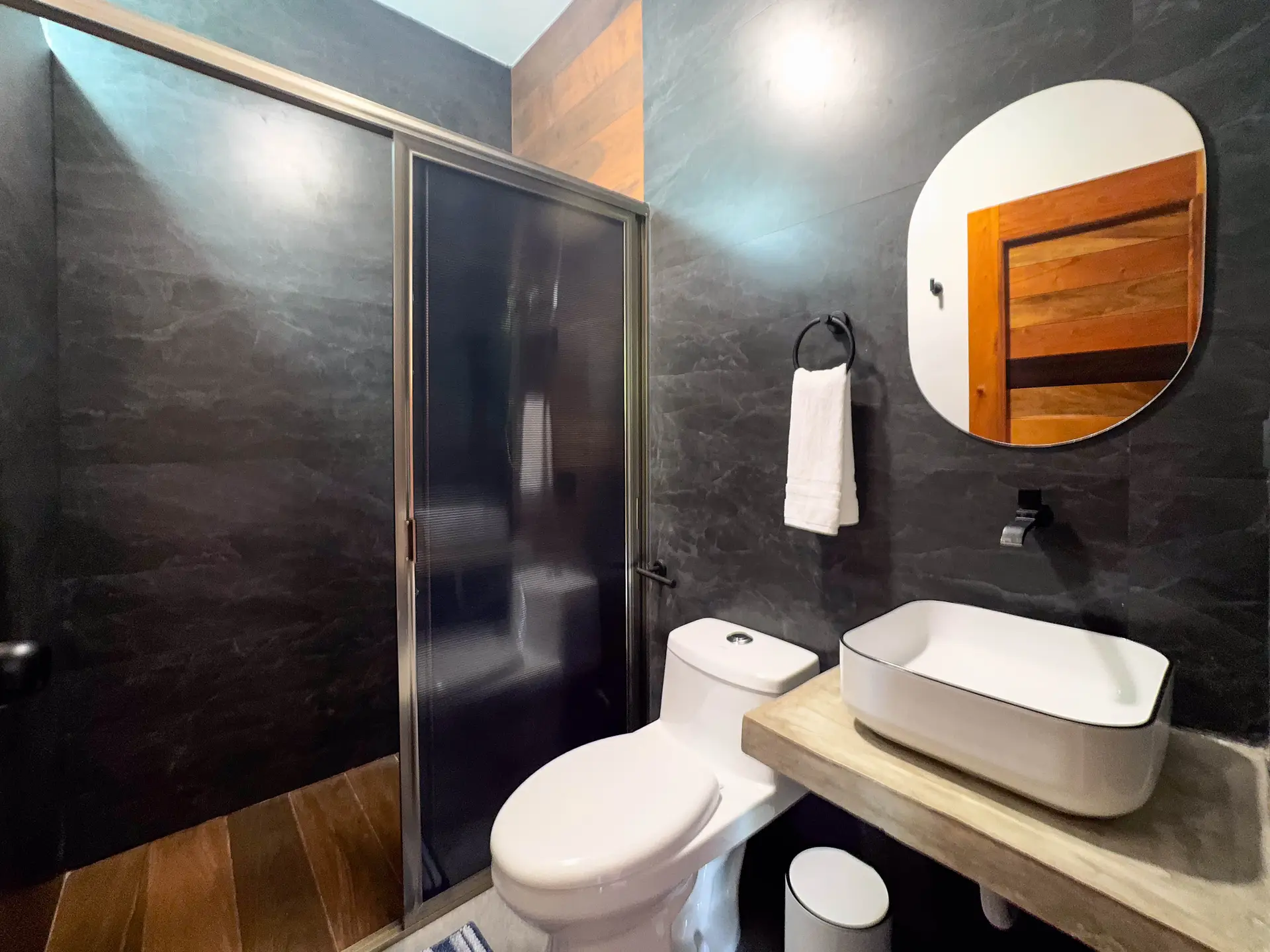 Baño de azulejo oscuro con ducha de cancel, lavabo de vasija y espejo redondo