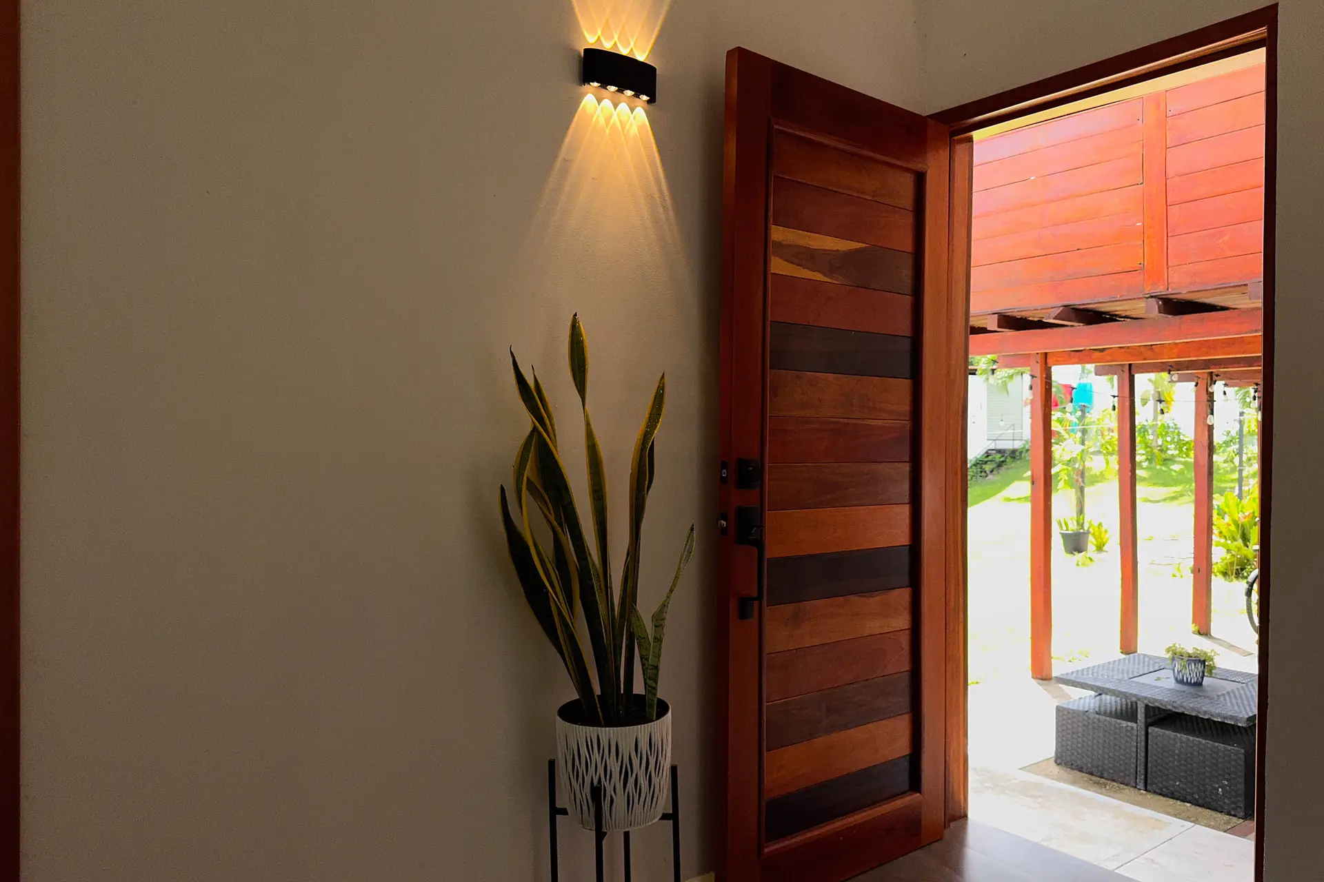 Puerta de madera abierta hacia el corredor exterior, con una planta y una lámpara de pared