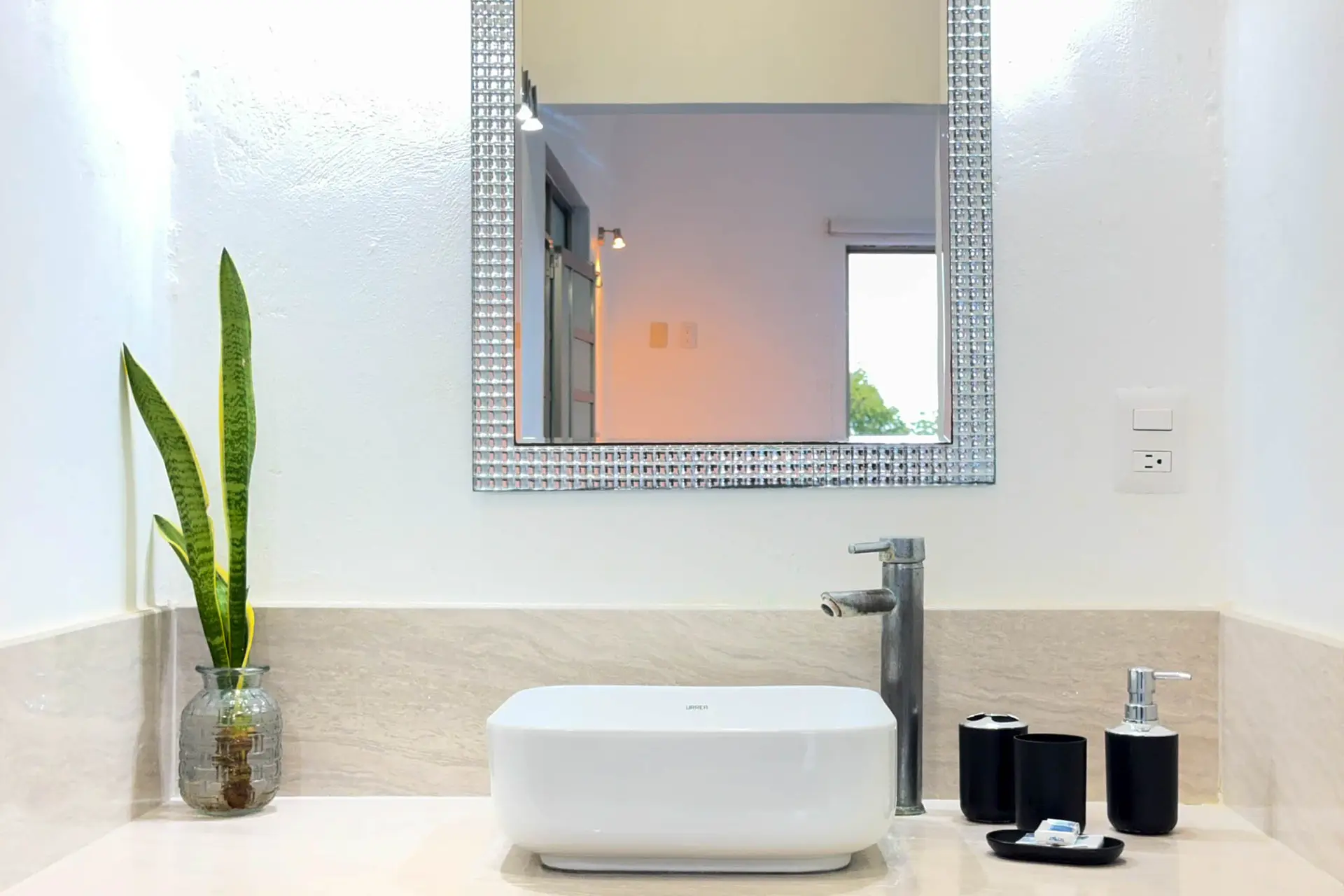 Lavabo de vasija blanca bajo un espejo de marco de mosaico, con una planta al lado