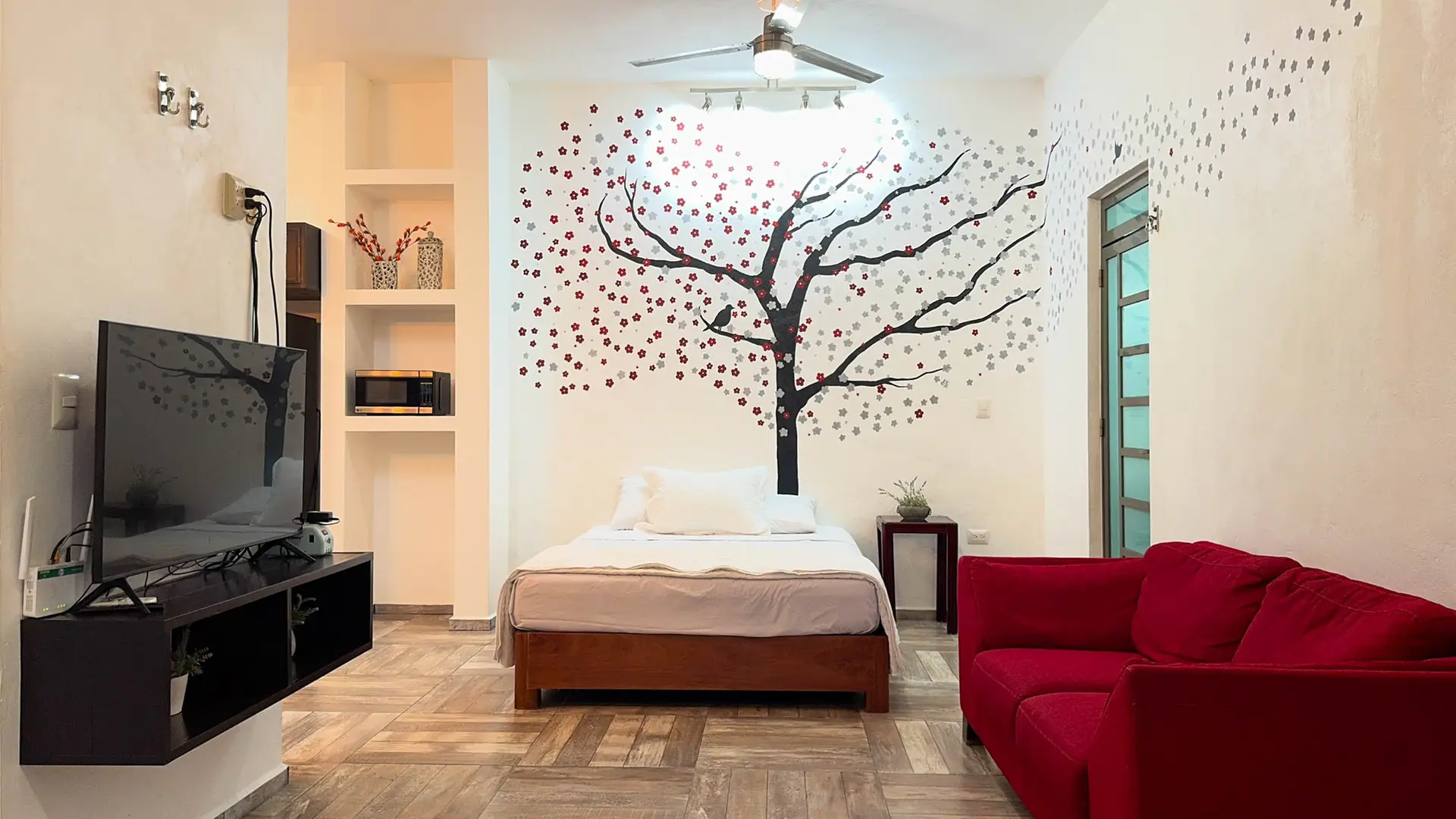 Cama individual bajo un árbol pintado en la pared, con televisión y un sofá rojo al lado
